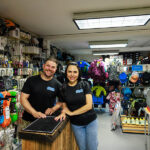 A man and a woman stand shoulder to shoulder against a backdrop of outdoor equipment.