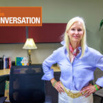 Vernier Science Education CEO Jill Hedrick, wearing a blue button-down shirt and a scarf, stands in front of a desk.
